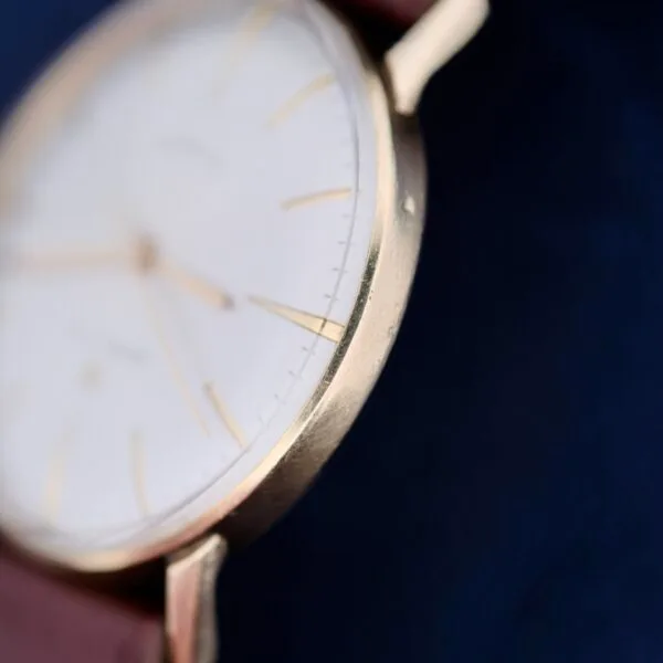 A close-up of a 1960s Hamilton Automatic Sunburst Dial wristwatch shows slight wear on the gold case, adding character. The watch has a white face with thin gold hour markers against a deep blue background. The brown leather strap is partially visible while the time remains indistinct.