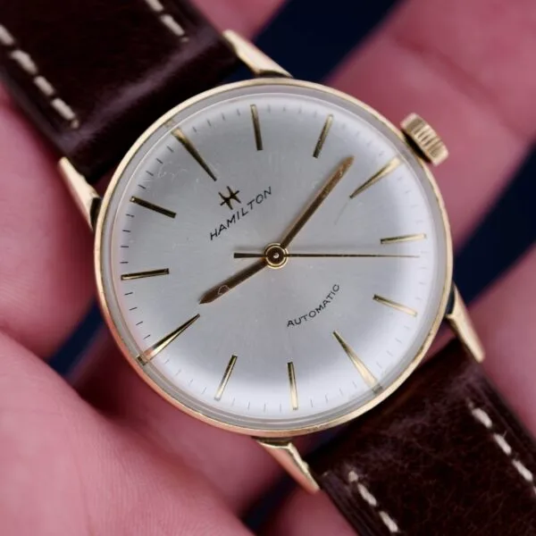 A close-up of a vintage 1960s Hamilton Automatic Sunburst Dial watch shows its silver dial, gold hour markers and hands, and classic brown leather strap with visible stitching. The brand name is centered on the dial as its held in a persons hand.