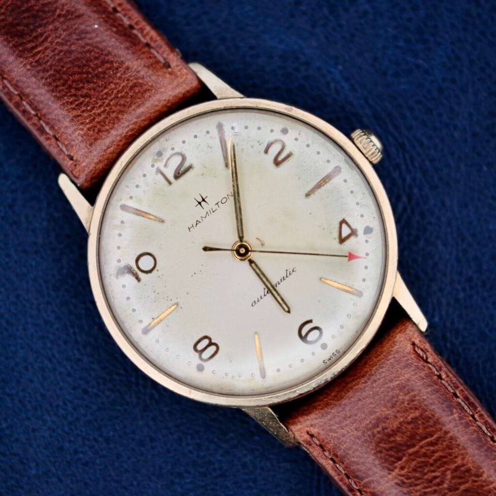 A 1950s Hamilton Automatic wristwatch with a brown leather strap is displayed against a blue background. It boasts a round face, gold-tone casing, simple hour markers, gold hands, and an elegant slender red-tipped second hand.