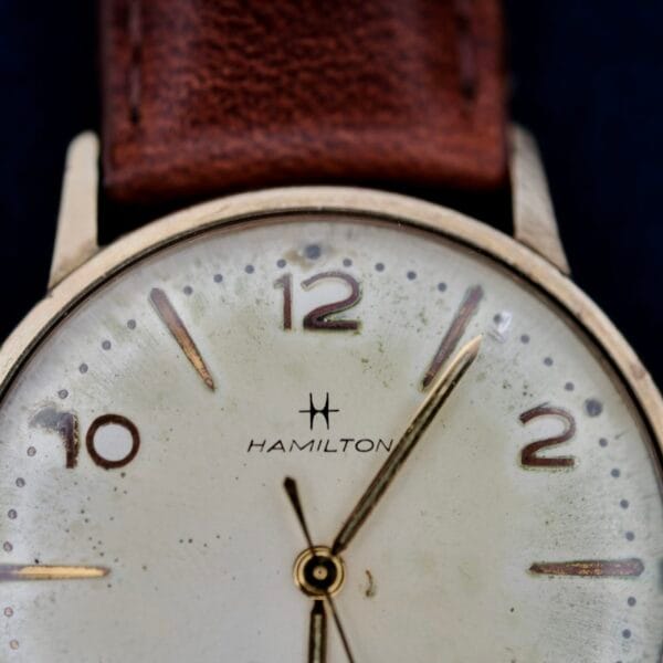 Close-up of a 1950s Hamilton Automatic wristwatch, featuring a round face with black numerals at 12, 2, 4, 8, and 10. Thin hour and minute hands accompany a small seconds hand on an aged face with a brown leather strap.