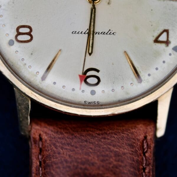 A close-up of the 1950s Hamilton Automatic wristwatch reveals a cream face with elegant gold hour markers. Numbers 8, 6, and 4 stand out, with a small red arrow near the number 6. It features a classic brown leather strap and subtly inscribed automatic on the face.