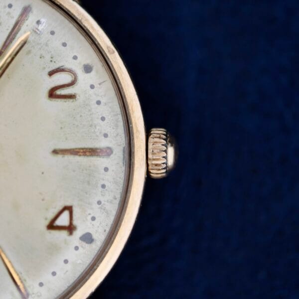 A close-up of a 1950s Hamilton Automatic wristwatch face highlights the numbers 2 and 4. The metallic edge gleams with a side crown against a deep blue background, exuding aged charm with its weathered appearance and visible speckles.