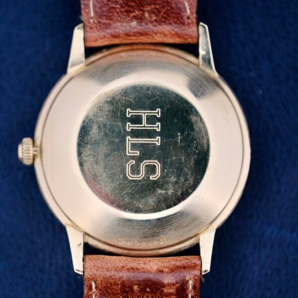 The image displays a 1950s Hamilton Automatic vintage wristwatch with a brown leather strap. The metallic back is engraved with SHS, enhancing its charm. Set against a dark blue background, the watch proudly shows some earned scratches.