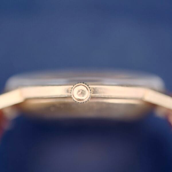 Side view of a 1950s Hamilton Automatic watch crown with textured edges, set on a gold case against a blurred blue backdrop. The centered crown, gold lugs, and partial face radiate timeless elegance.
