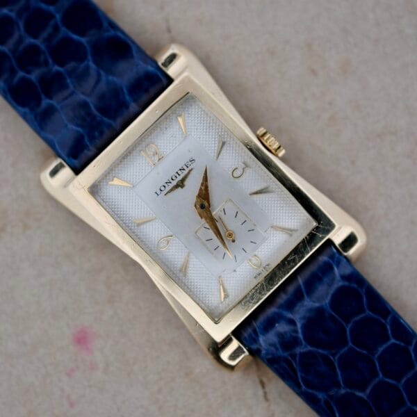 The 1950s Longines “Hour Glass” Case watch features a rectangular gold-toned design, white dial with black minute and hour hands, gold hour markers, a small seconds sub-dial at 6 oclock, and is paired with a blue textured strap.