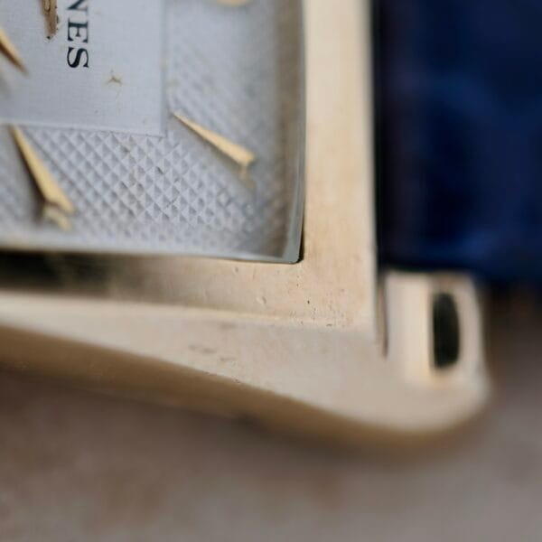 Close-up of a 1950s Longines “Hour Glass” gold wristwatch corner, showcasing a textured geometric dial with gold Roman numerals. The edge of a blue strap is visible in the background, with NES featured on the watch face.