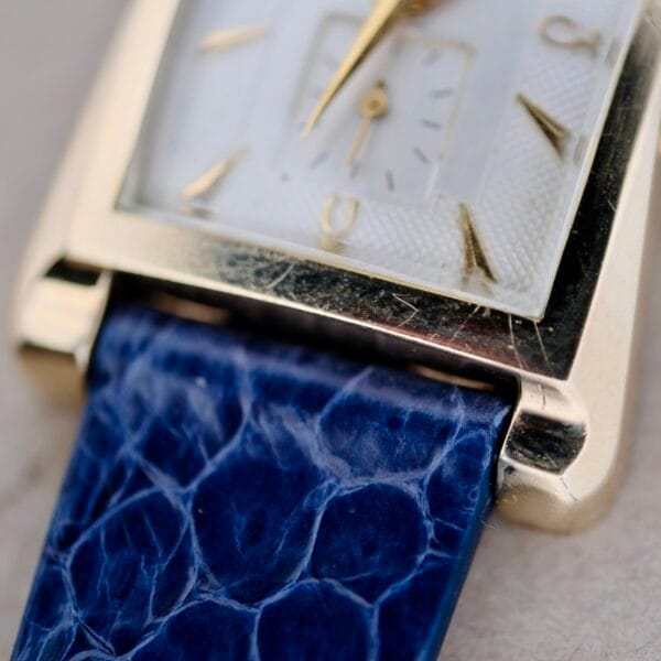 The 1950s Longines “Hour Glass” wristwatch showcases a gold frame with a white face, featuring gold hour markers and hands. Its complement is a blue textured leather strap, all set against a soft-focus neutral background.