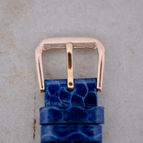 Close-up of the 1950s Longines “Hour Glass” case featuring a metallic, gold-finished buckle on a textured blue strap against a light grey background.