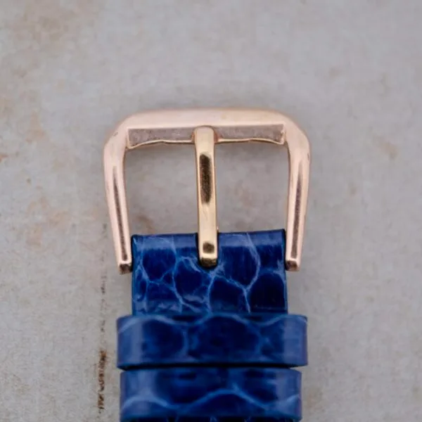 Close-up of the 1950s Longines “Hour Glass” case featuring a metallic, gold-finished buckle on a textured blue strap against a light grey background.