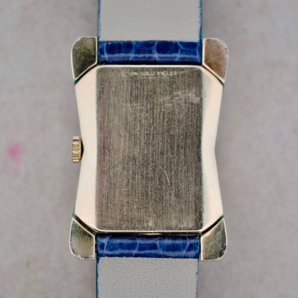 A close-up of the back of the 1950s Longines “Hour Glass” case shows a rectangular, gold-toned wristwatch with a textured surface and a small crown on the left. It features a strap made from blue and white textured material.
