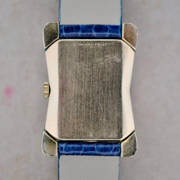A close-up of the back of the 1950s Longines “Hour Glass” case shows a rectangular, gold-toned wristwatch with a textured surface and a small crown on the left. It features a strap made from blue and white textured material.