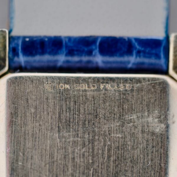 Close-up image of a 1950s Longines “Hour Glass” Case, showcasing a textured, worn metallic surface inscribed with 10K GOLD FILLED. A worn blue textured band borders the text.
