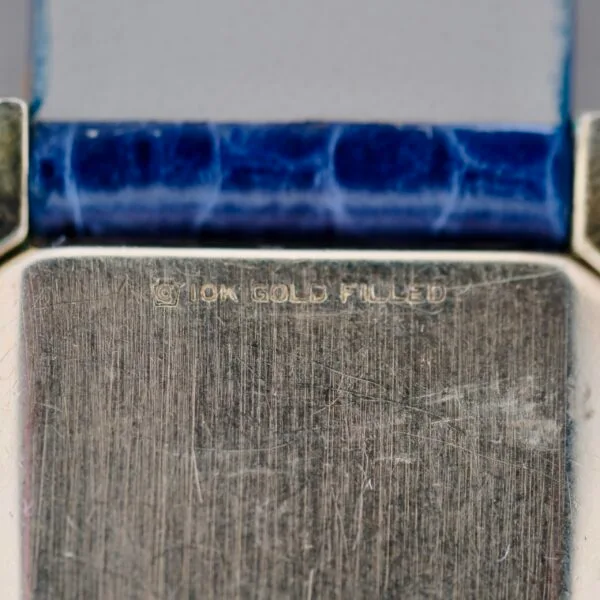 Close-up image of a 1950s Longines “Hour Glass” Case, showcasing a textured, worn metallic surface inscribed with 10K GOLD FILLED. A worn blue textured band borders the text.