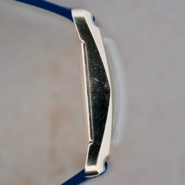 Close-up of a 1950s Longines “Hour Glass” wristwatch featuring a silver metal casing with visible scratches and a blue strap. The out-of-focus background highlights the watchs details.