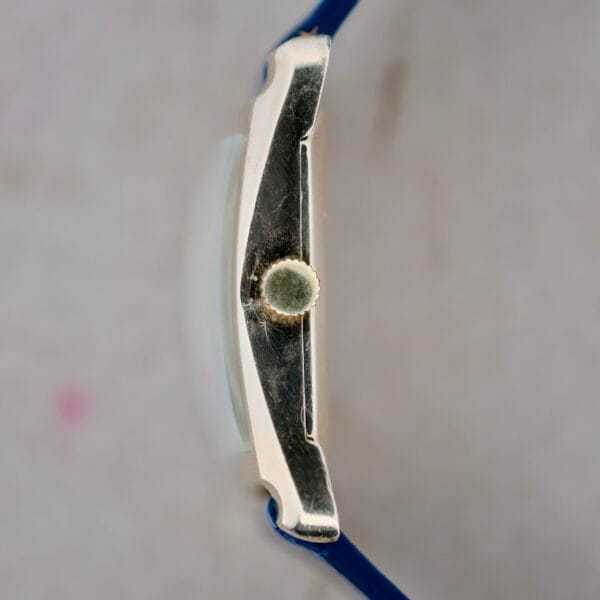 Side view of a 1950s Longines Hour Glass watch with a blue strap and silver case. The crown for setting the time is visible in the center, set against a blurred neutral background.