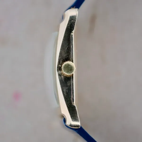 Side view of a 1950s Longines Hour Glass watch with a blue strap and silver case. The crown for setting the time is visible in the center, set against a blurred neutral background.