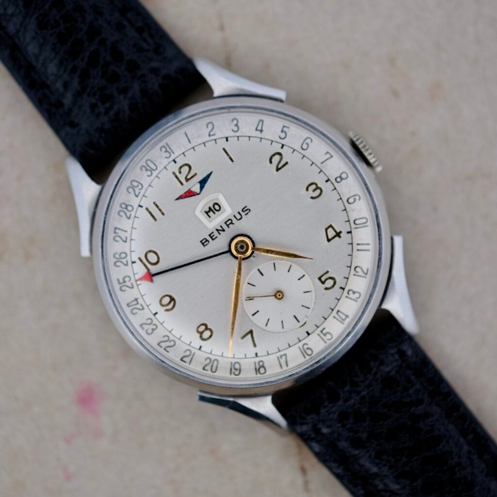 A Benrus Triple Calendar in Steel, featuring a silver dial, gold hands, date display, and black strap is shown on a light surface.