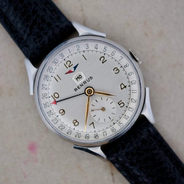 Close-up of a Benrus Triple Calendar In Steel wristwatch with a black leather strap, showcasing a white dial, silver case, gold hands, date window at 12 oclock, and small seconds subdial above 6 oclock, resting on a light surface.