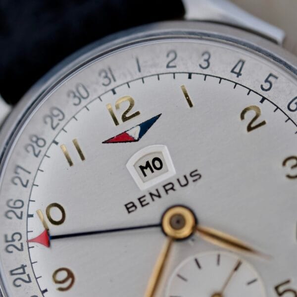 Close-up of a vintage Benrus Triple Calendar In Steel wristwatch. Features include gold hour markers and hands, an outer-edge date indicator, a central month display, a tachymeter scale, and a seconds subdial.