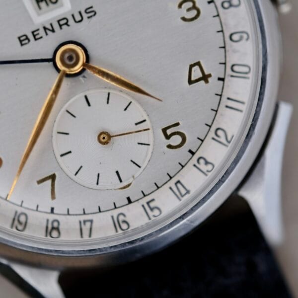 Close-up of the Benrus Triple Calendar In Steel watch face, showcasing gold hour/minute hands, a small seconds subdial at 6 oclock, black hour markers, and a date indicator around the edge. The silver-toned case has a partially visible leather strap.