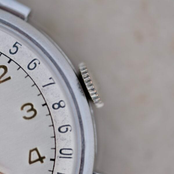 Close-up of the Benrus Triple Calendar in Steel, highlighting part of the dial with black numerals 2-8 and a grooved crown on the watch case. The background features a blurred neutral surface.