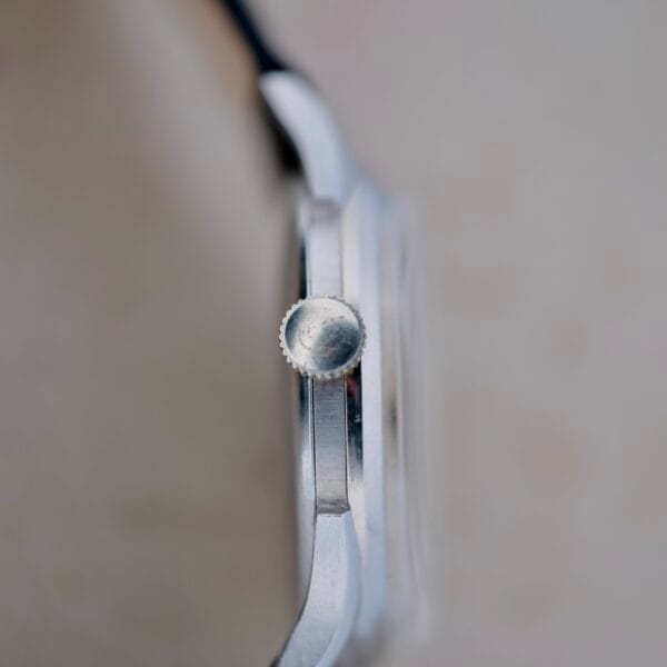 A close-up side view of the Benrus Triple Calendar In Steel watch case, featuring the crown on the right. The metallic case has a smooth finish, with a slightly textured edge. The blurred background enhances focus on this silver timepiece.