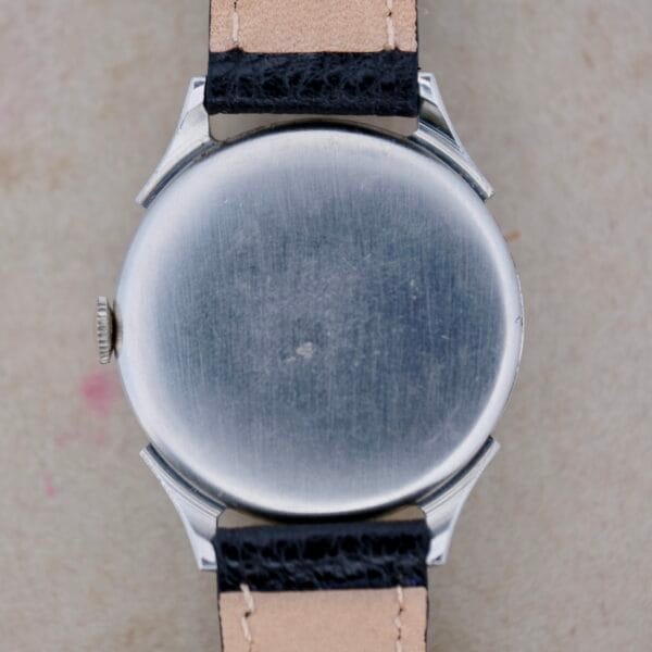The image features the back of the Benrus Triple Calendar In Steel wristwatch with a lightly brushed silver finish. It includes black leather straps accented with visible beige stitching and edges, set against a textured, light-colored surface.