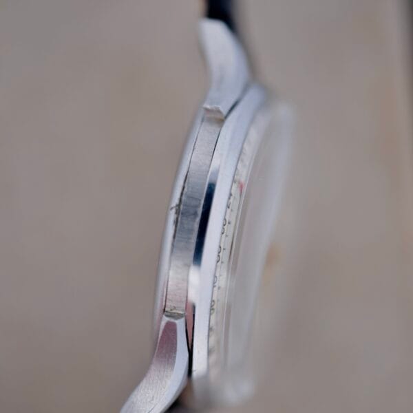 Close-up side view of the Benrus Triple Calendar In Steel watch case, highlighting the edge and part of the crown. The metallic surface and contours are in focus, while its face is slightly blurred against a soft, neutral background.