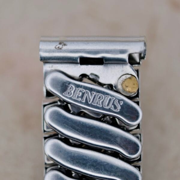 A close-up of the metallic watchband clasp on the Benrus Triple Calendar in Steel displays the engraved name Benrus, with a silver finish and a gold-toned component at the top right corner. The background remains blurred and neutral.