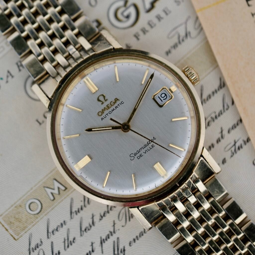 The Omega Seamaster Deville 166.020 watch features a silver dial and gold-tone hour markers, with the date on the 19th. It has a metal link band, shown in close-up resting on vintage paper with partial text visible.