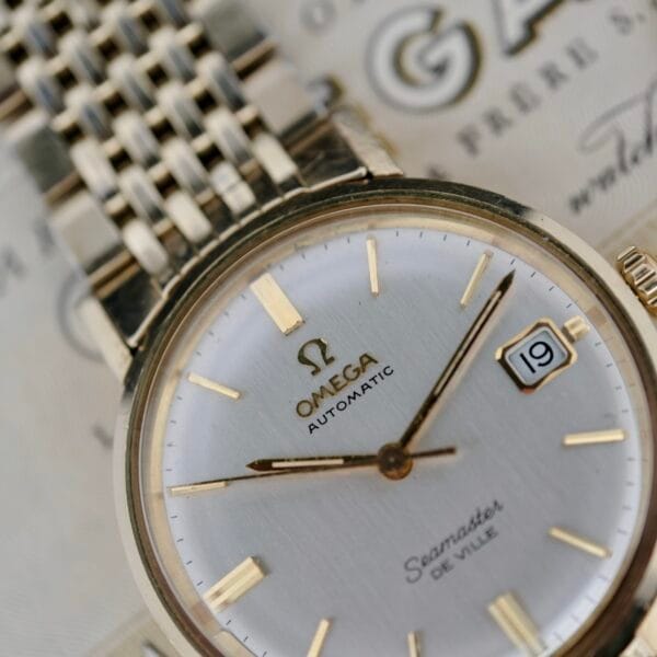 Close-up of the Omega Seamaster Deville 166.020 watch with a silver dial, gold markers and hands, showing the date 19 on a two-tone metal link bracelet against a blurred logo and text background.