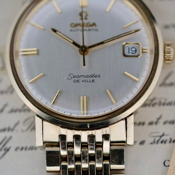 A detailed view of the Omega Seamaster Deville 166.020 Full Set watch, featuring a gold case, silver face with date set to 19th, and metal bracelet, placed on paper with cursive writing.