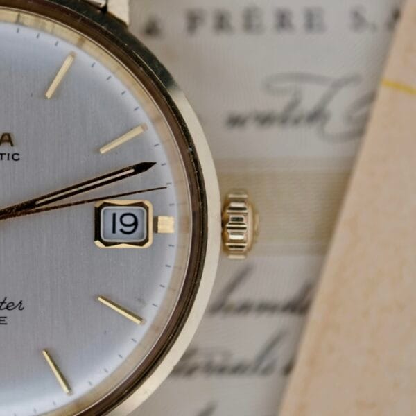 A close-up of the Omega Seamaster Deville 166.020 Full Set vintage wristwatch showcases its silver-toned face with gold markers. The date, 19, is visible in a small window against a textured background featuring cursive writing in muted colors.