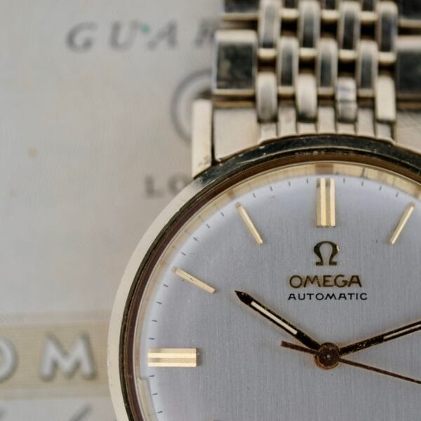 Close-up of an Omega Seamaster Deville 166.020 wristwatch featuring a silver dial, gold hour markers, and a matching bracelet with hands indicating approximately 10:10. The blurred background displays the Omega logo.