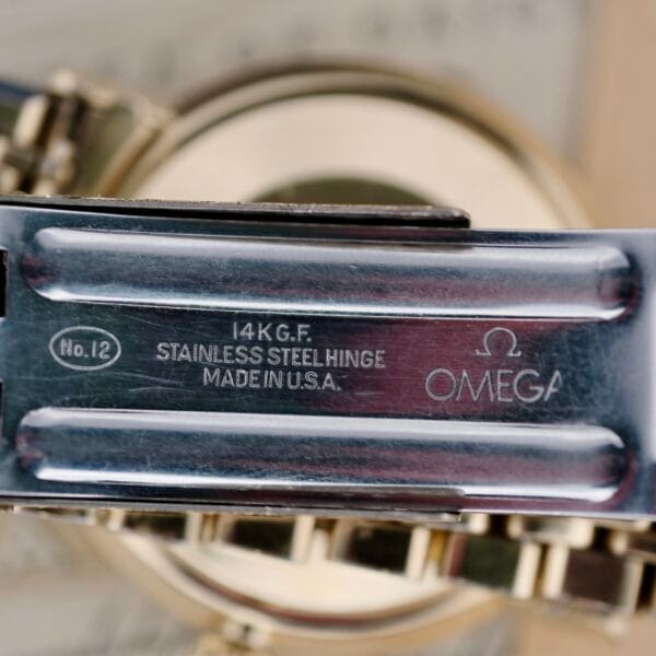 Close-up of the Omega Seamaster Deville 166.020 clasp, featuring 14K G.F., STAINLESS STEEL HINGE, MADE IN U.S.A., No.12, and the Omega logo inscriptions on a polished metallic background. The setting is blurred for emphasis.