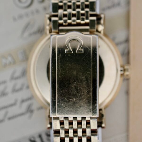 A close-up of the Omega Seamaster Deville 166.020 Full Set wristwatchs metal clasp bearing the Omega logo, with a blurred background of documents. The woven metal bracelet shows signs of wear, including visible scratches on the clasp surface.