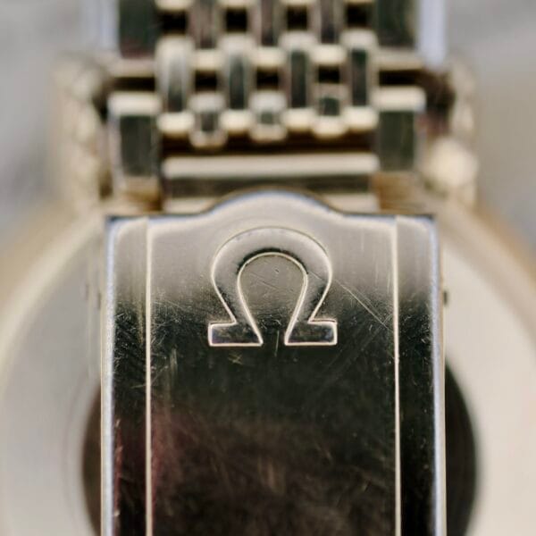 Close-up of the Omega Seamaster Deville 166.020s metal watch clasp, showcasing an engraved Omega symbol on its brushed surface, connecting to a woven metal strap.