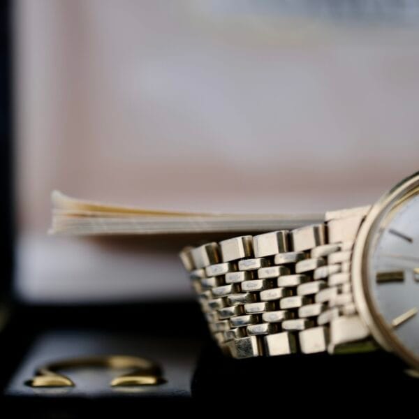 A close-up of the Omega Seamaster Deville 166.020 Full Set displays its gold-toned wristwatch with a metallic band on a dark surface, featuring a minimally detailed watch face. In the background, theres a blurred book and an indistinct rectangular object.