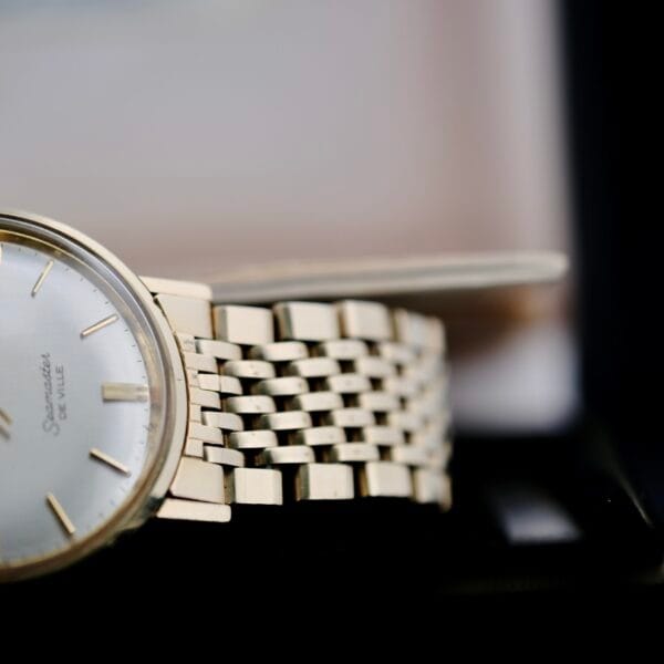 Close-up of an Omega Seamaster Deville 166.020 watch with a metallic band and light face. It features thin hands, small hour markers, and part of the brand name visible, set against a softly blurred background.