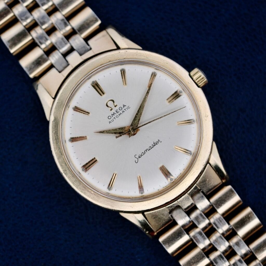 An Omega Seamaster L6298 Dauphine Hands wristwatch with a gold-toned metal bracelet is set on a dark blue surface. It features a white round face, gold hour markers and hands, along with the classic Omega Automatic and Seamaster inscriptions on the dial.