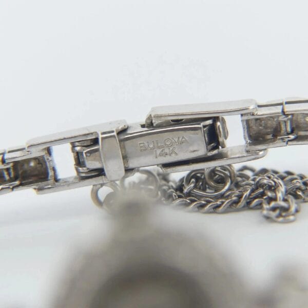 Close-up view of a silver wristwatch clasp engraved with BULOVA 14K from a 1950s Bulova 14K Ladies White Gold & Diamond Watch, attached to a metal chain. The focus is on the clasp with part of the chain visible in the foreground.