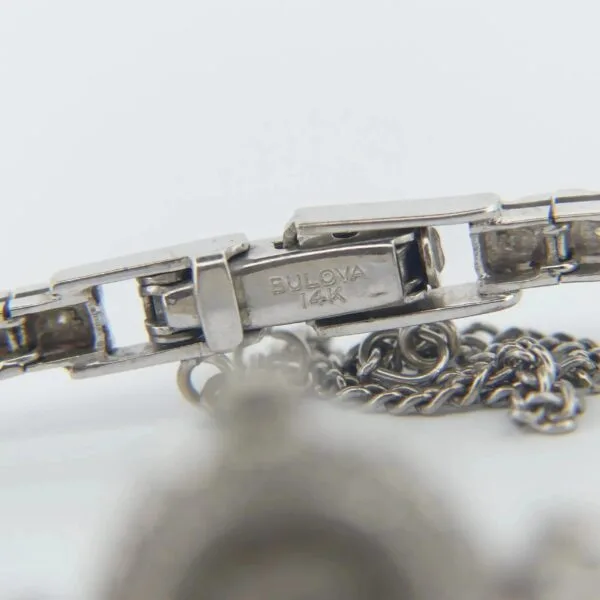 Close-up view of a silver wristwatch clasp engraved with BULOVA 14K from a 1950s Bulova 14K Ladies White Gold & Diamond Watch, attached to a metal chain. The focus is on the clasp with part of the chain visible in the foreground.