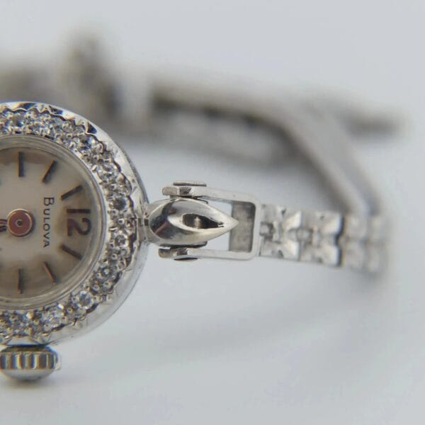 Close-up of a 1950s Bulova 14K Ladies White Gold & Diamond Watch with a bejeweled bezel and metal strap, displaying the brand name, silver hour markers, and ornate design. The time shows around 4:50.