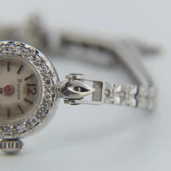 Close-up of a 1950s Bulova 14K Ladies White Gold & Diamond Watch with a bejeweled bezel and metal strap, displaying the brand name, silver hour markers, and ornate design. The time shows around 4:50.