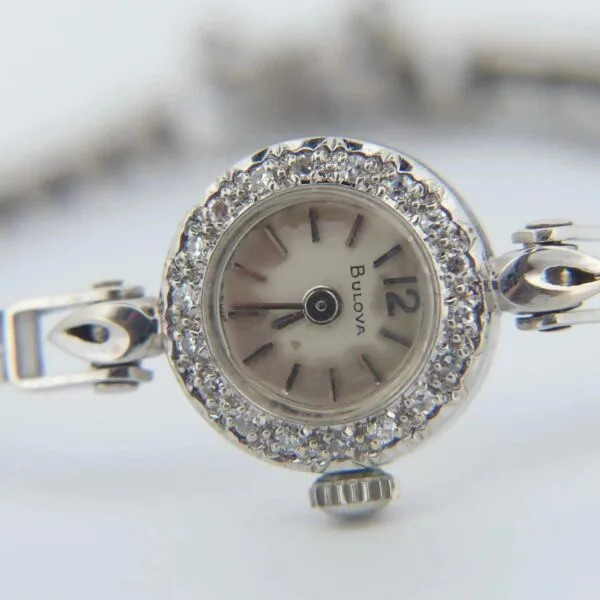Close-up of a vintage 1950s Bulova ladies wristwatch, crafted in 14K white gold with diamonds. It features a silver metal band, a white face with black hour markers and hands, and a decorative textured bezel. The number 12 is prominently displayed.