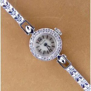 A 1950s Bulova 14K ladies white gold watch features a small round face encircled by clear stones, black hands and numerals on a light dial, and an intricately floral-designed metal strap, all set against a tan surface.