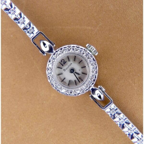 A 1950s Bulova 14K ladies white gold watch features a small round face encircled by clear stones, black hands and numerals on a light dial, and an intricately floral-designed metal strap, all set against a tan surface.