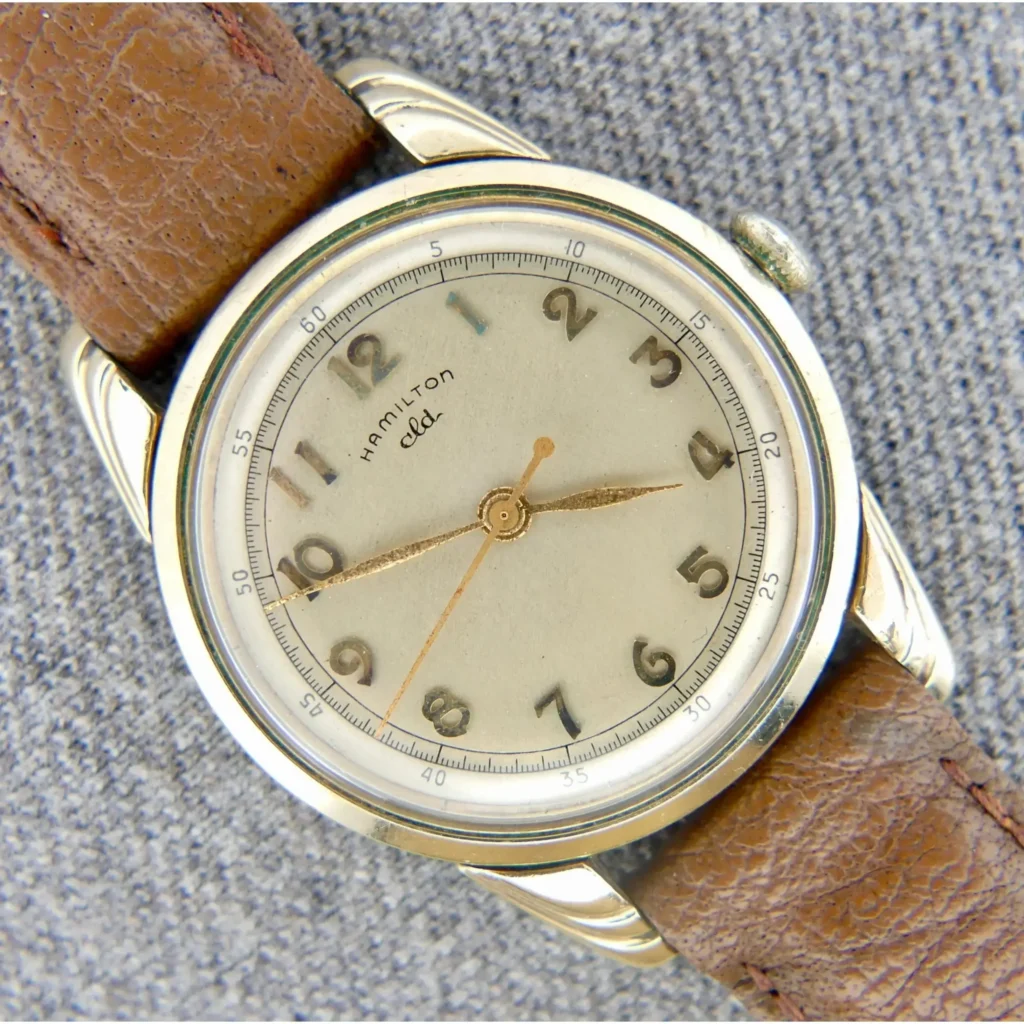 A 1950s Hamilton Lyndon CLD Stepped Lug Gold Filled wristwatch with a round case and brown leather strap, cream dial with Arabic numerals, and gold hands, displayed on a gray textured fabric background.