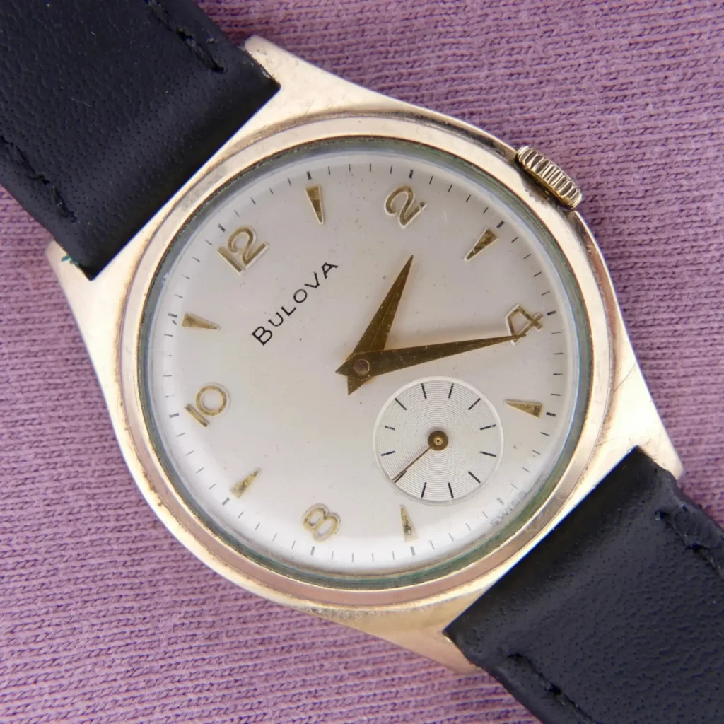 A 1959 Bulova Senator Cal. 11AF wristwatch with a white dial, gold numerals and hands, seconds subdial, and black leather strap rests on a textured purple surface.