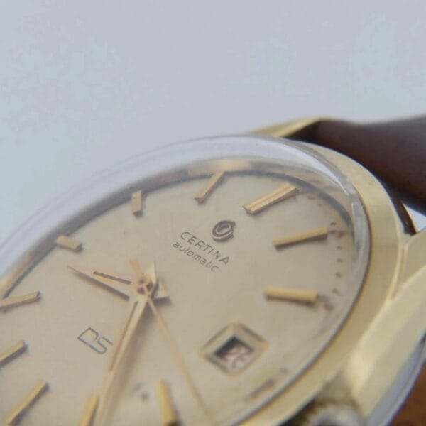 Close-up of a 1959 Certina DS Automatic 1st Gen. wristwatch featuring a beige dial, gold hour markers and hands. It has a 14k gold-toned case with a brown leather strap and includes a small date window.
